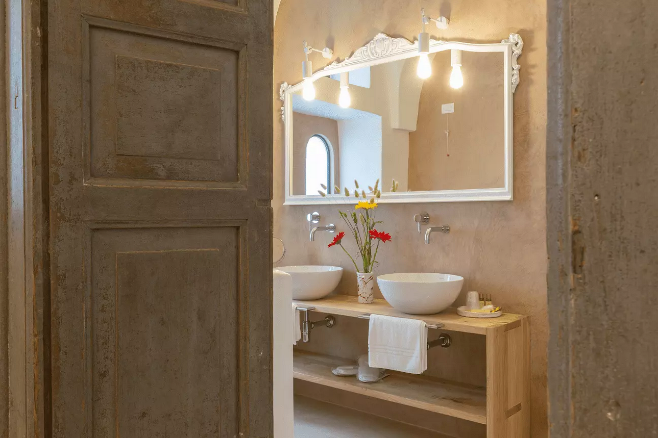 Esposizione di sanitari da bagno con rubinetti, lavandini e mensole su una parete grigia e una colonna di legno.