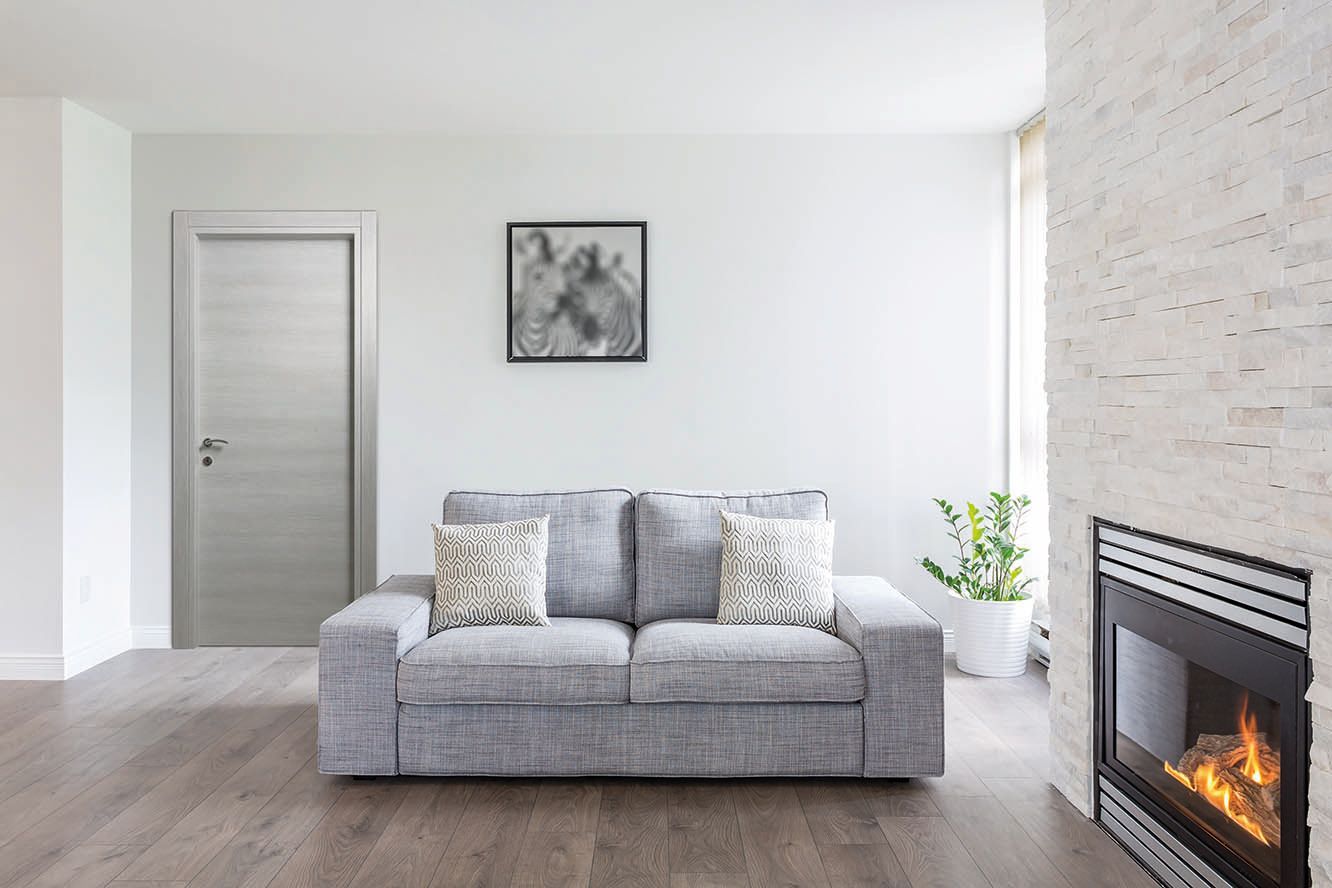 Un divano grigio è posizionato di fronte a una parete bianca, una porta e un camino con una fiamma accesa.