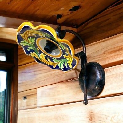 Applique decorativa con paralume in ceramica con motivi floreali blu, gialli e verdi, montata su una parete in legno.
