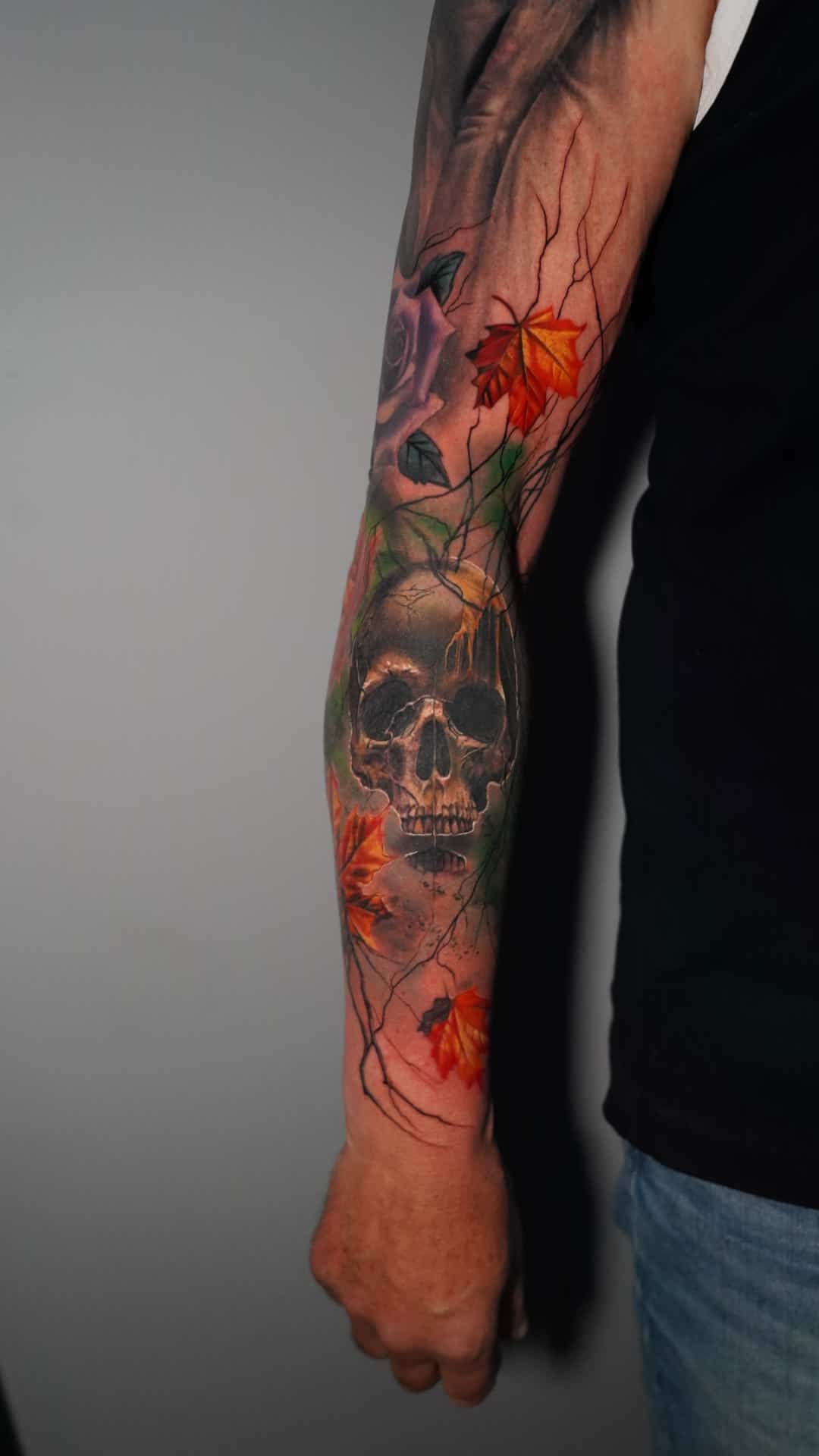 An arm sleeve tattoo featuring a central human skull surrounded by vibrant, colorful autumn maple leaves.