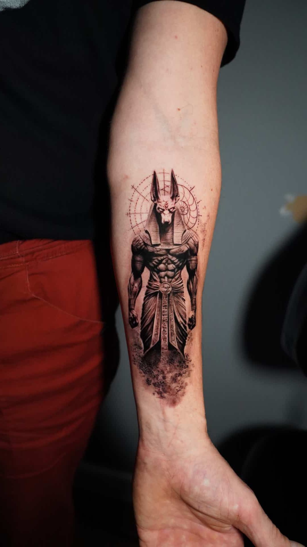 Detailed black and grey tattoo of the Egyptian god Anubis on a person's inner forearm, featuring geometric linework.