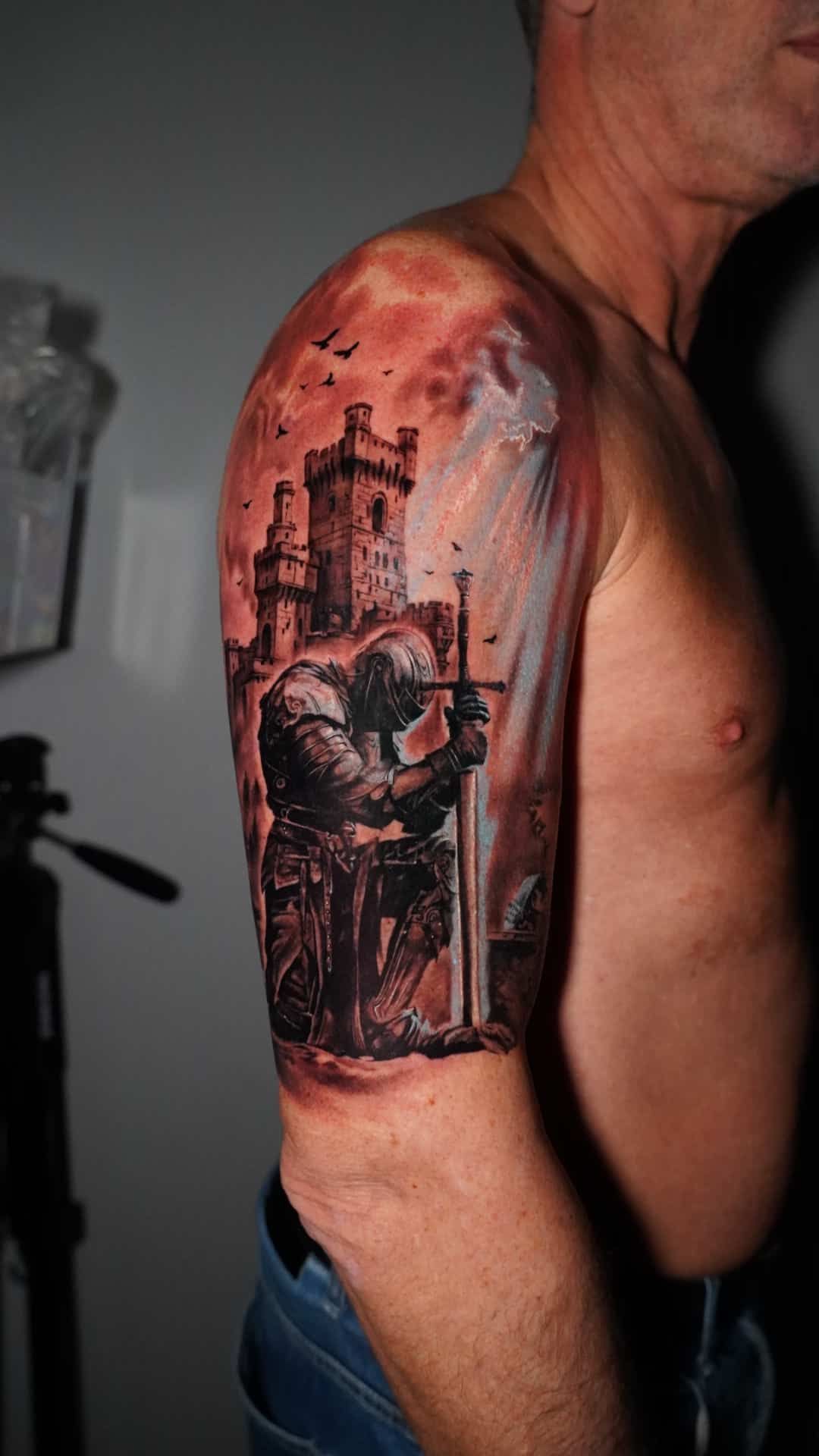 A black and red tattoo on a person's upper arm depicting a knight kneeling before a sword in front of a stone castle.