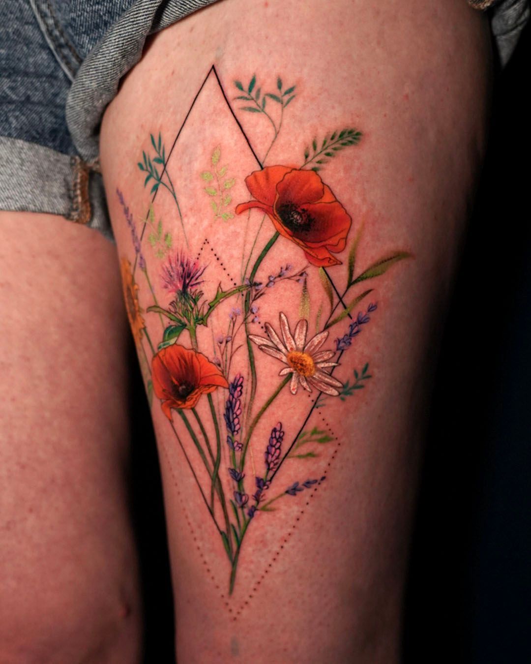 A botanical thigh tattoo featuring two red poppies, a daisy, and purple wildflowers within a geometric diamond frame.
