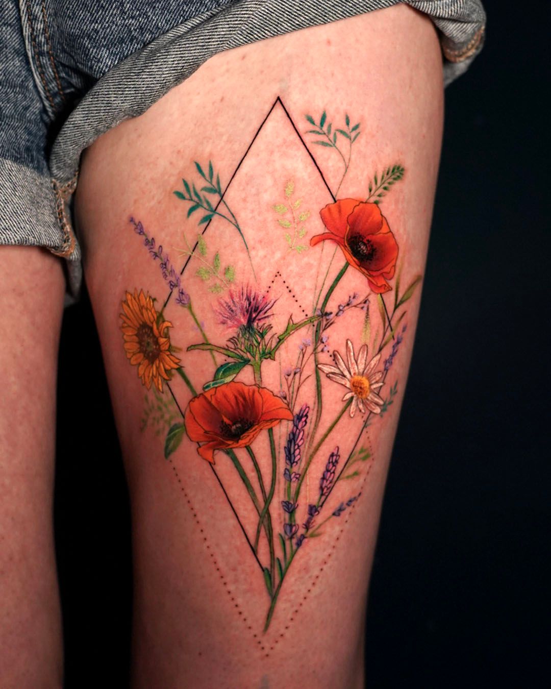 A vibrant floral tattoo featuring red poppies, a sunflower, and purple wildflowers contained within a diamond outline.