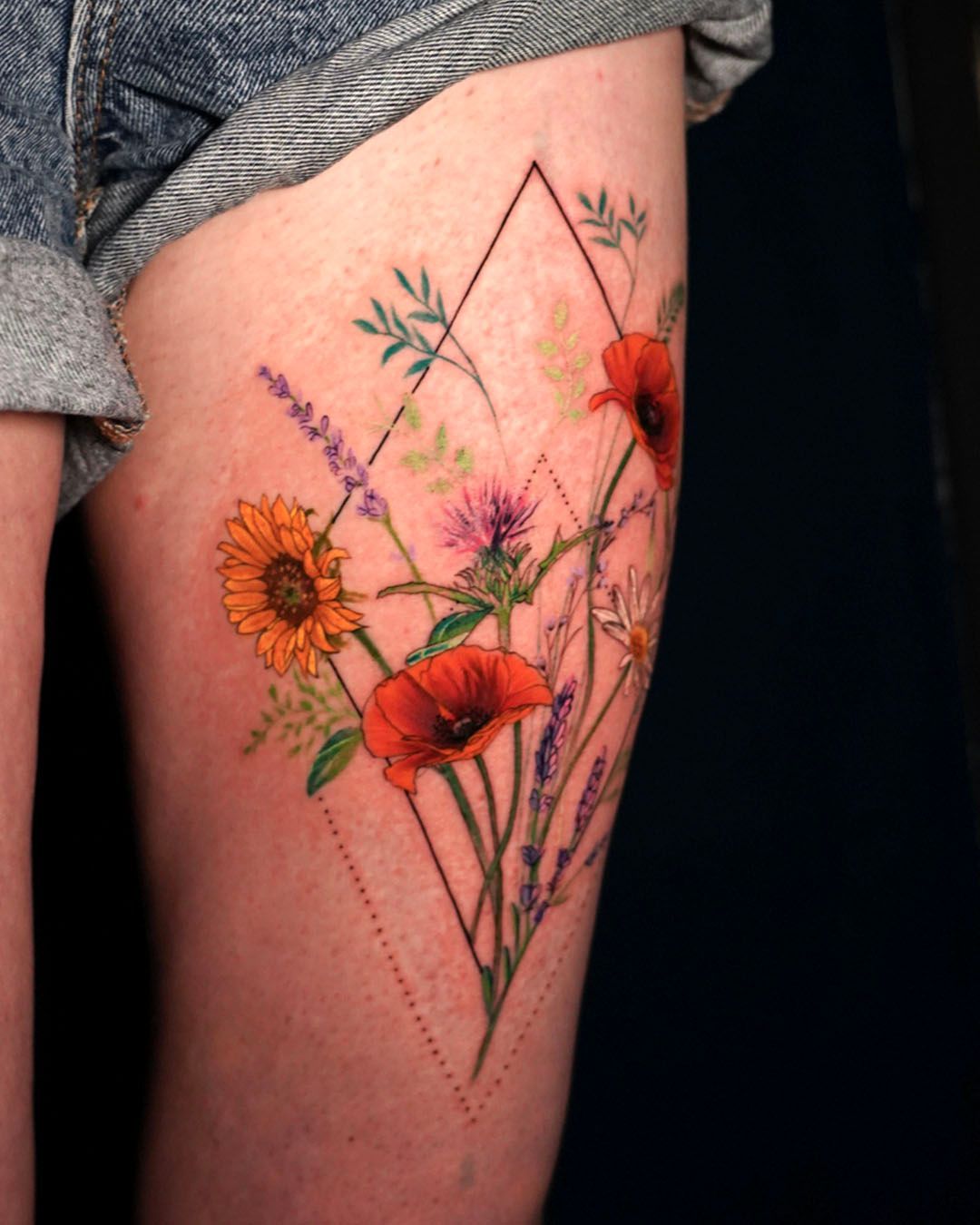 A colorful botanical thigh tattoo featuring wildflowers like poppies, lavender, and daisies framed by a geometric diamond.