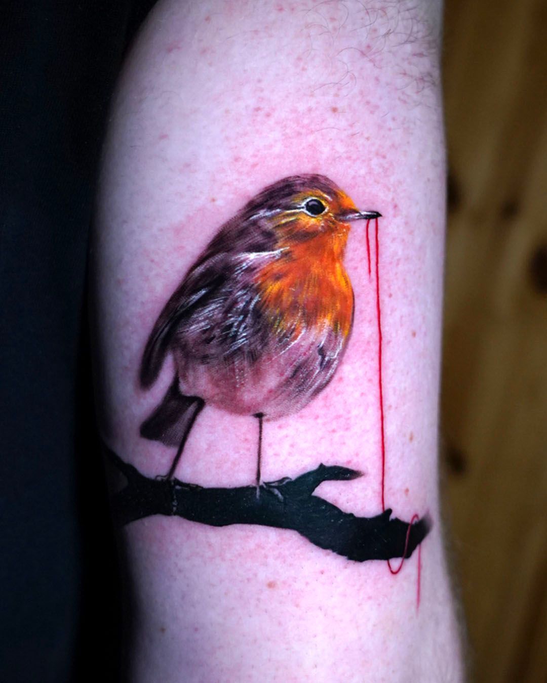 A realistic tattoo of a robin perched on a black branch, holding a long, trailing red thread in its beak.