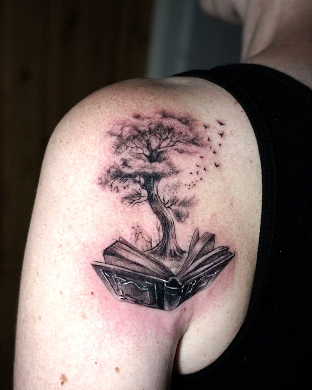 A shoulder tattoo featuring a black-and-grey ink design of a tree growing out of an open book, with small birds flying off.