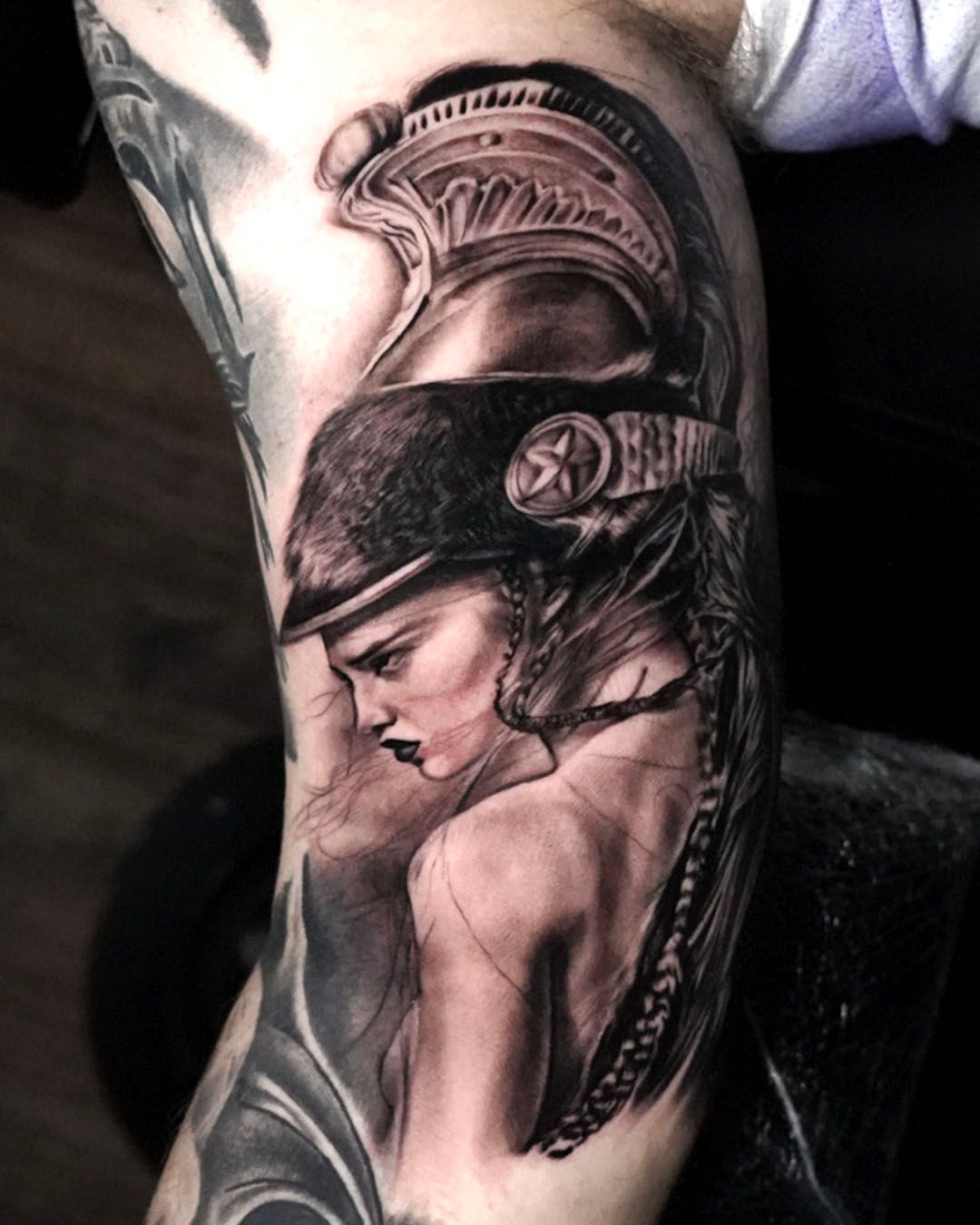 Black and grey tattoo of a warrior in a classic helmet, shown in profile with a stern expression and braided hair.