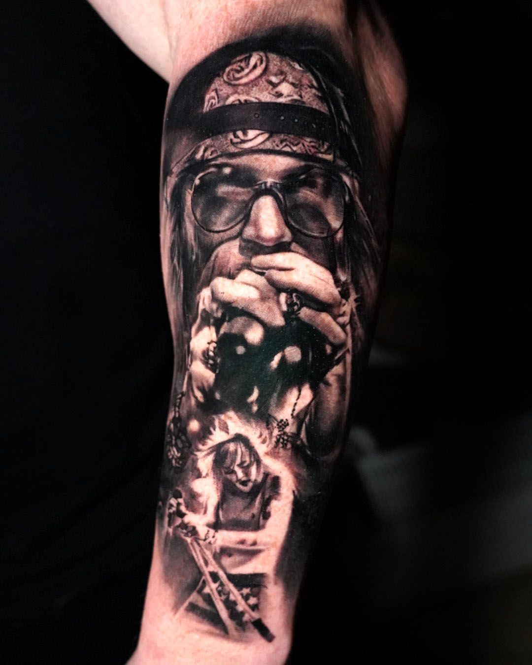 A black-and-grey tattoo of a person wearing a bandana and glasses, holding a microphone, with a smaller figure below.