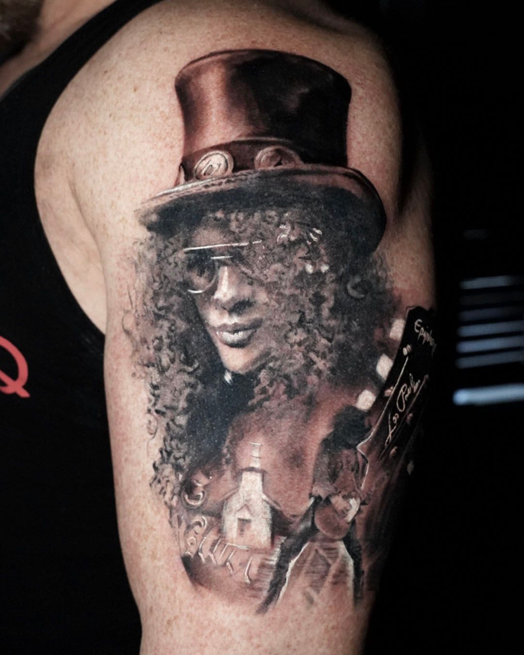 A realistic, monochromatic tattoo on an upper arm depicting Slash wearing his signature top hat and curly hair.