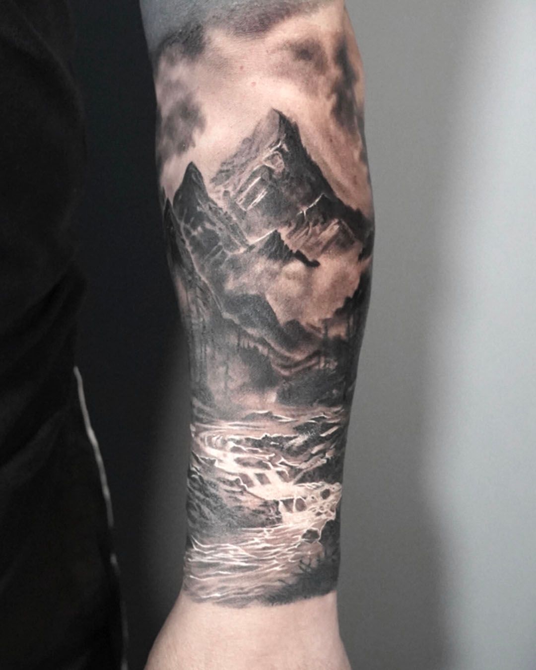 A black and grey tattoo on an arm featuring a detailed mountain range reflected in a flowing river under a cloudy sky.