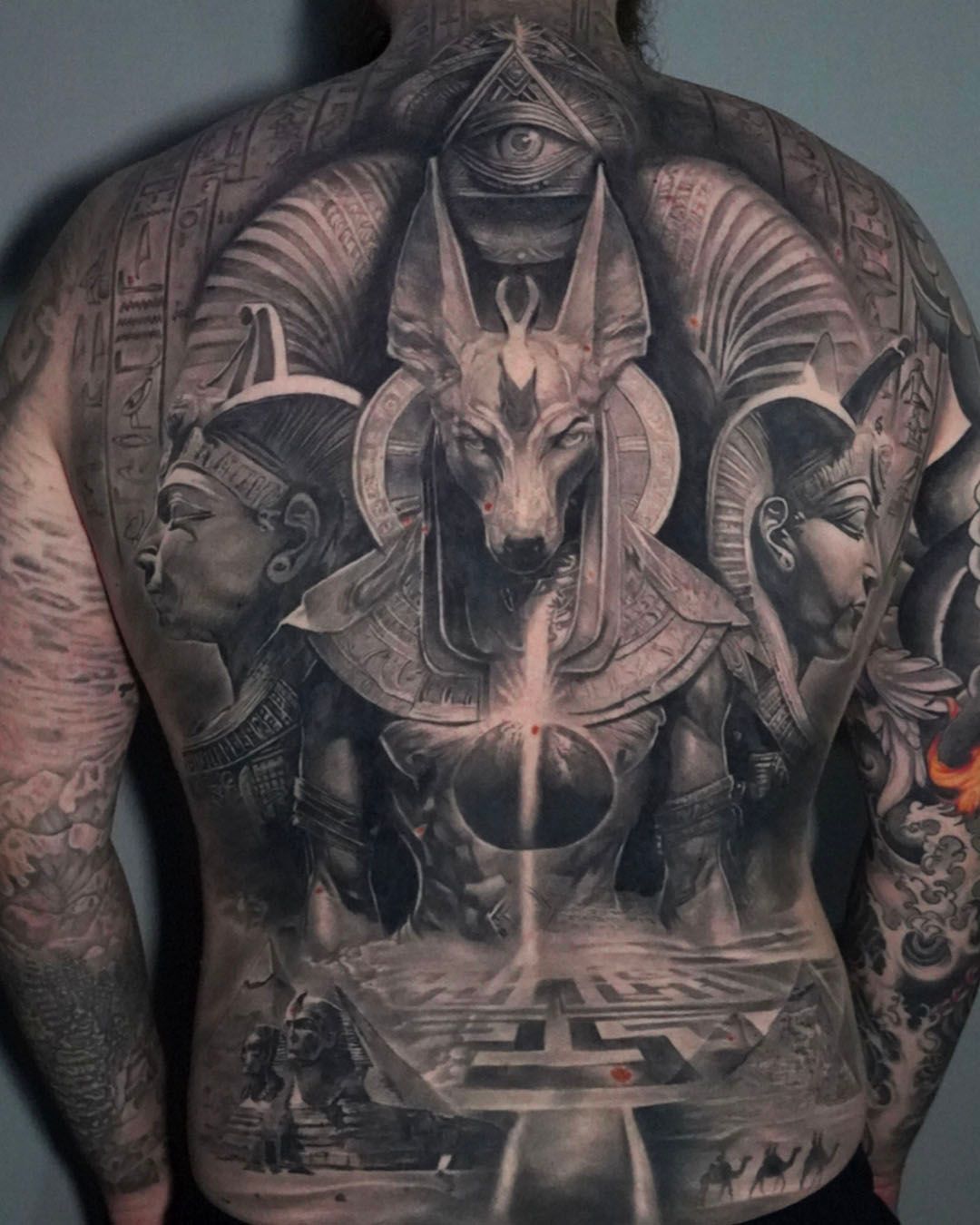 Black and grey back tattoo featuring a central Anubis figure flanked by Egyptian royalty under an all-seeing eye.