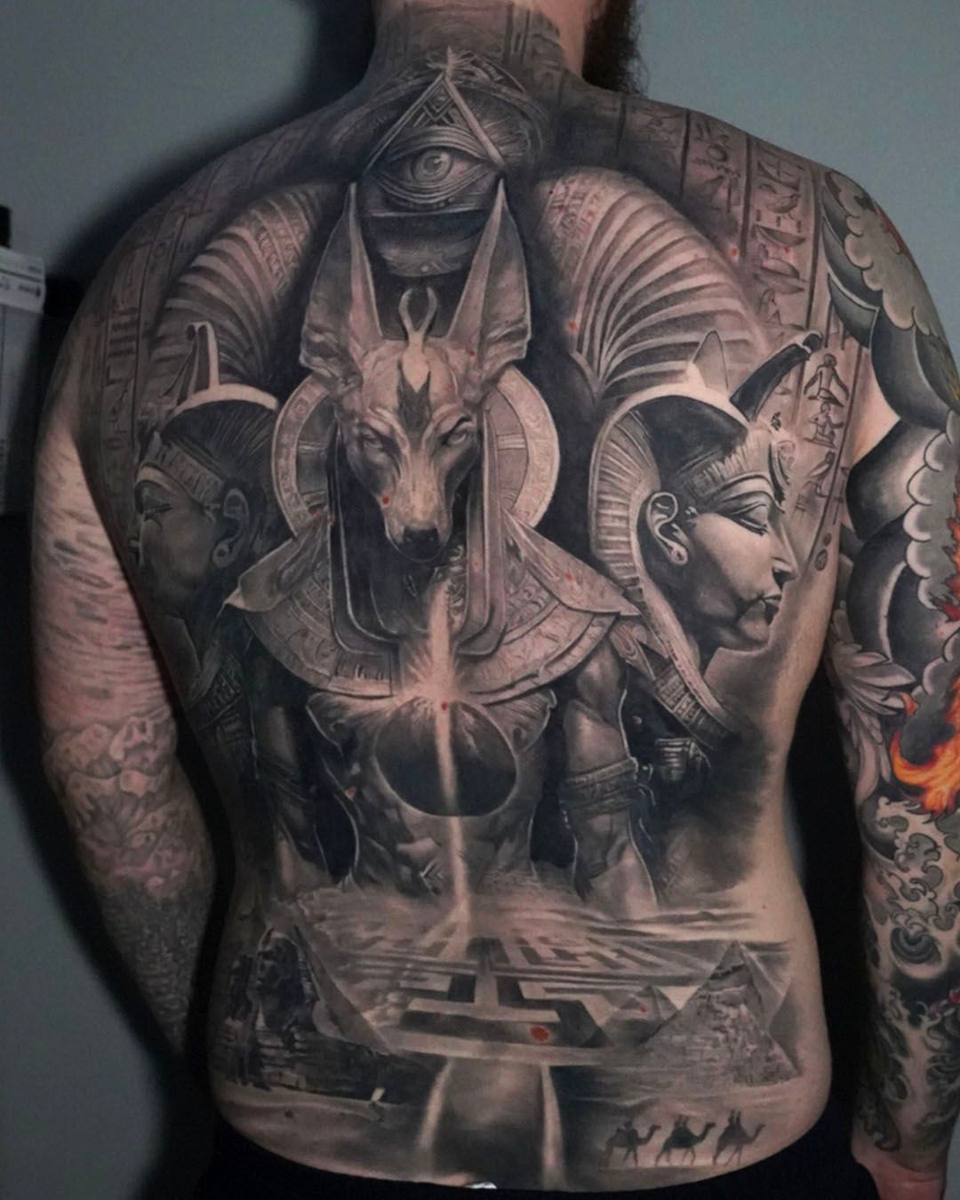 An intricate black-and-grey back tattoo featuring Anubis, Egyptian pharaohs, hieroglyphics, and pyramid iconography.