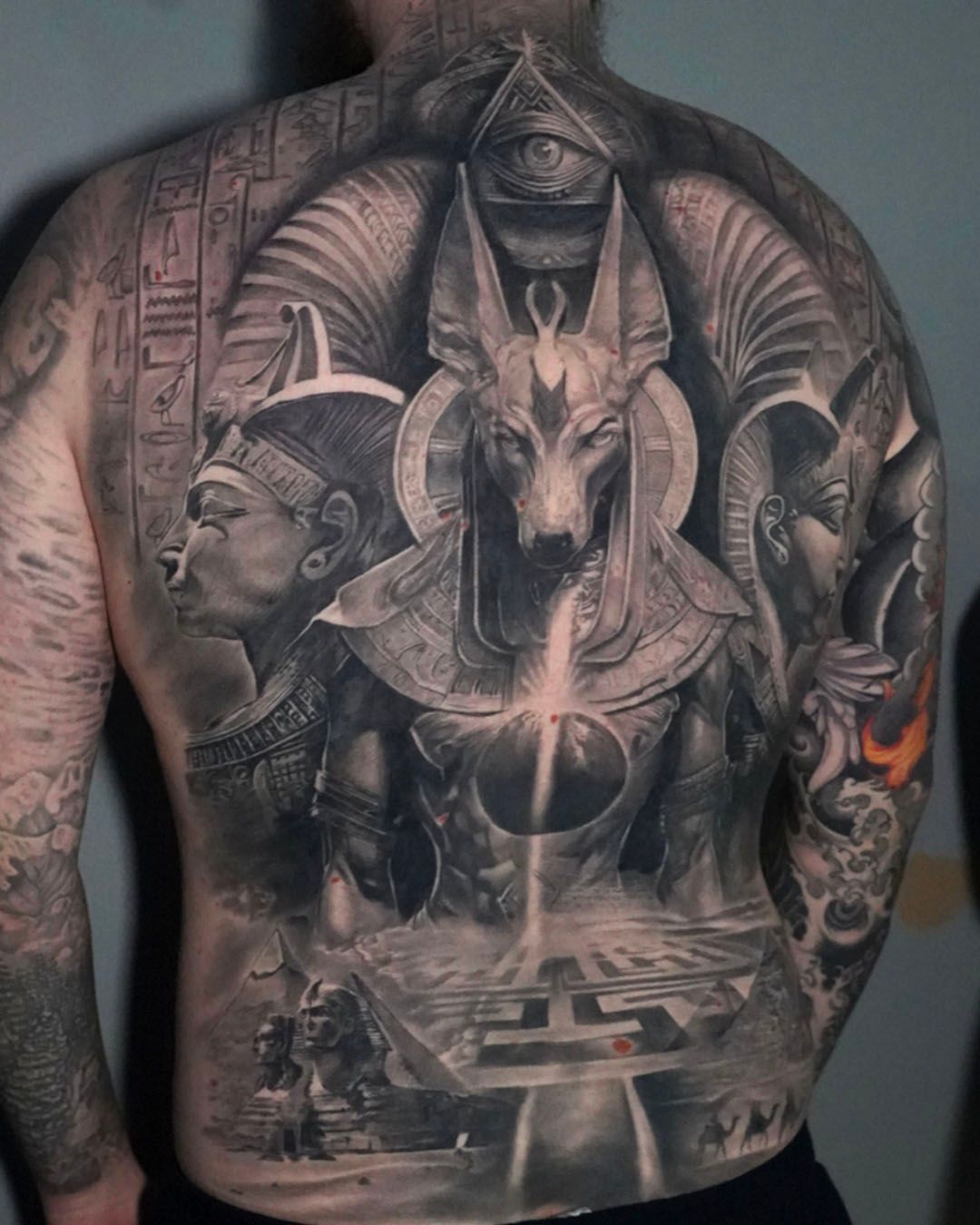A detailed black and grey back tattoo featuring Anubis, an Egyptian pharaoh, the Eye of Providence, and hieroglyphs.