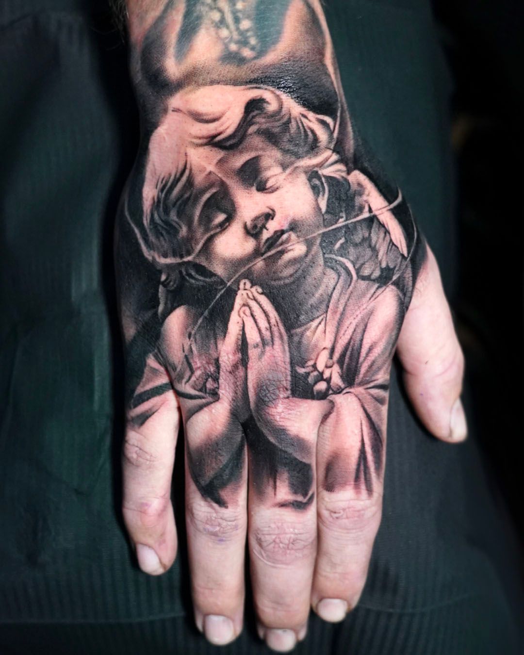 A black-and-grey tattoo on the back of a hand depicting a cherub with closed eyes in a praying pose.