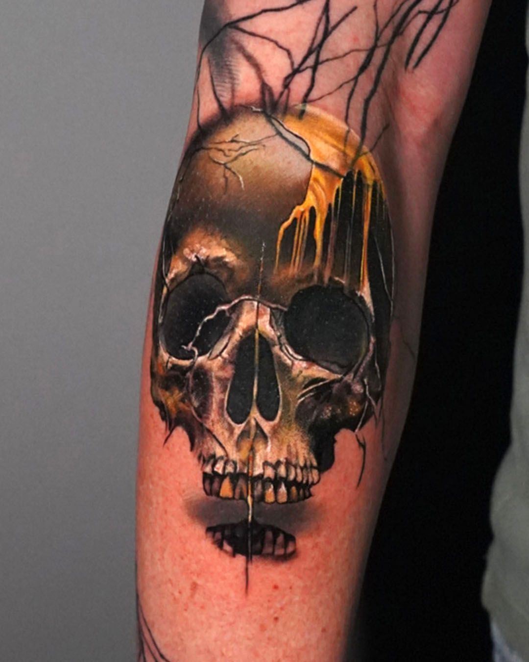 A realistic tattoo of a skull on an arm, with yellow liquid dripping over the top and black branches extending upward.