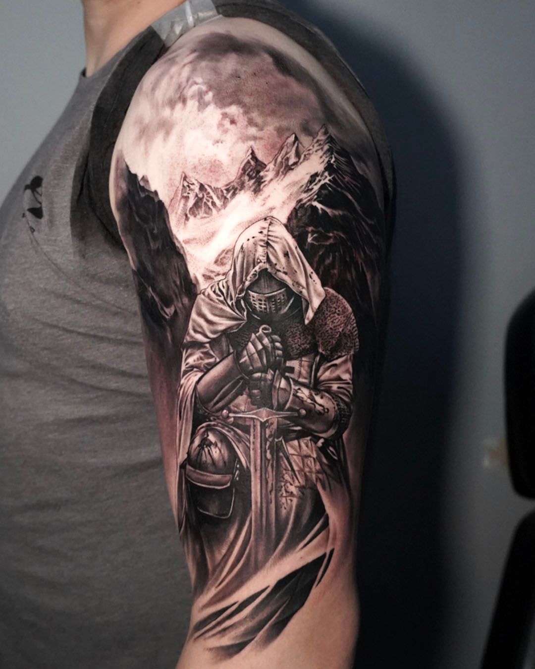 A black-and-grey tattoo on an upper arm depicting a knight in a hooded cloak kneeling before a sword, set against mountains.