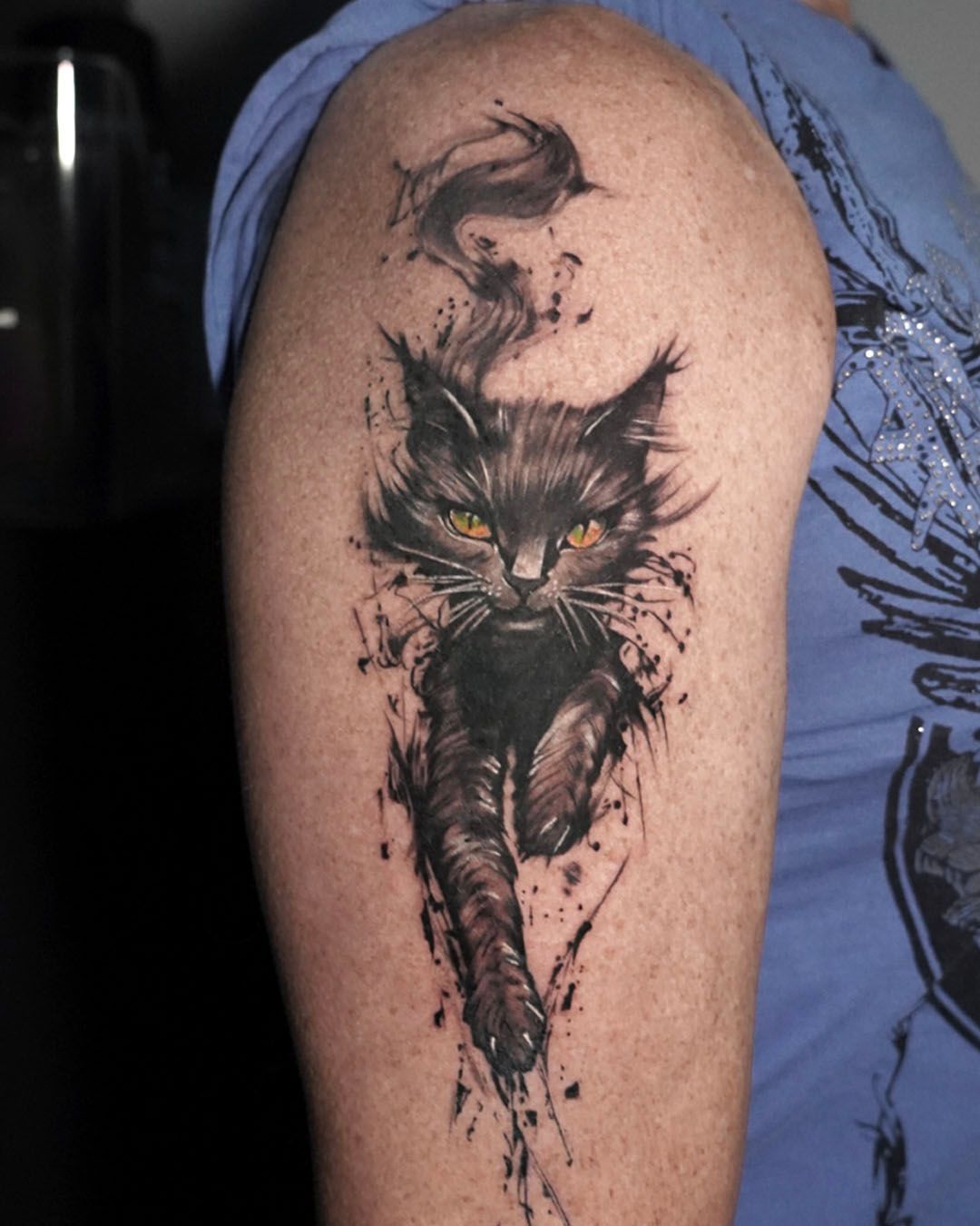 A tattoo on an upper arm featuring a black cat with striking amber eyes, depicted in a flowing, ink-splash art style.
