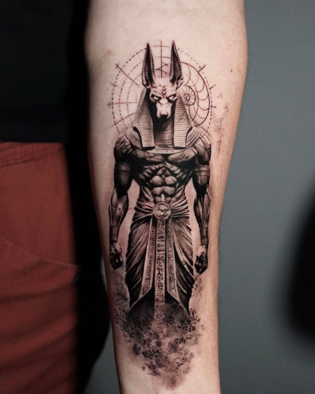 A black-and-grey tattoo on a forearm depicting the Egyptian god Anubis with geometric line work in the background.