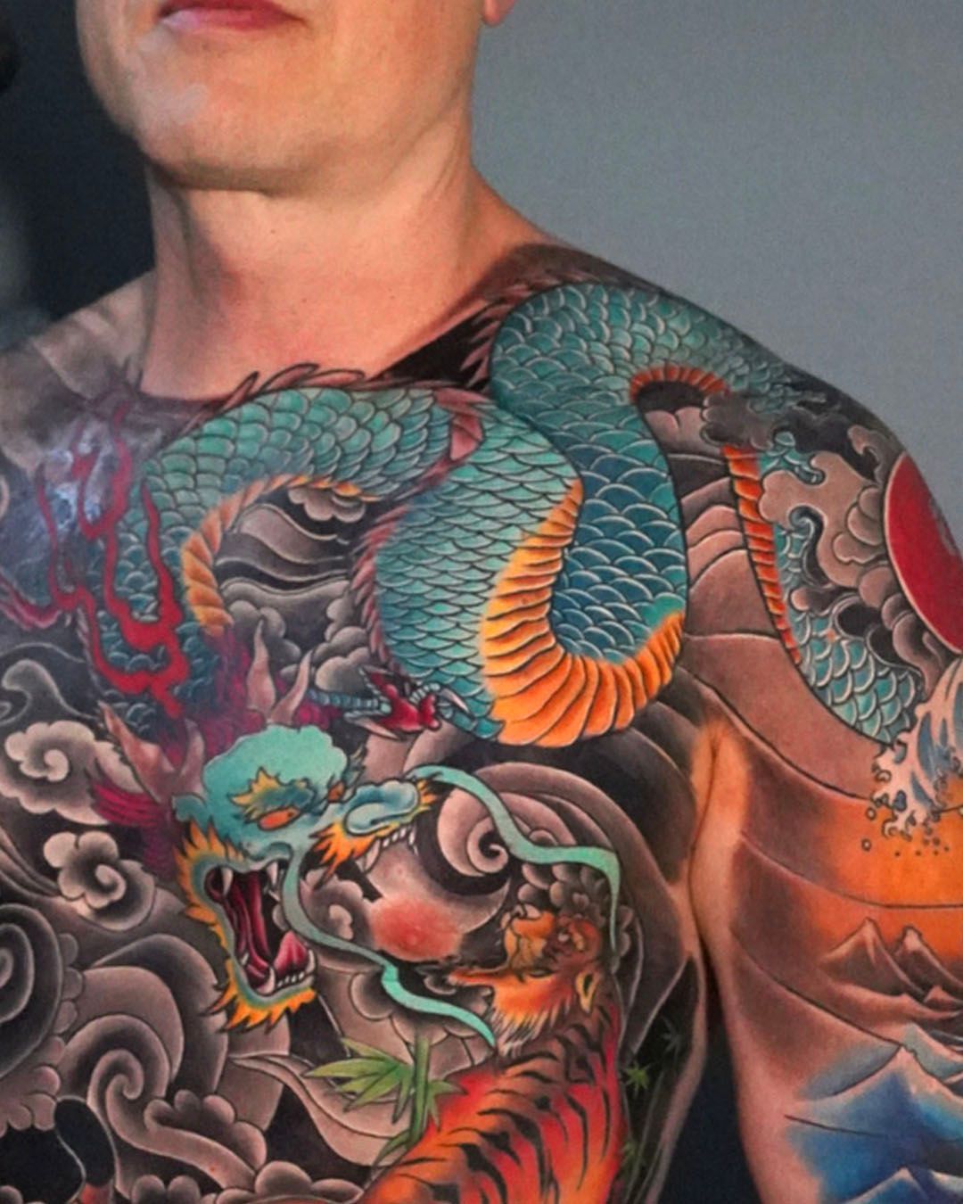 A man's chest and shoulder featuring an intricate, colorful Japanese-style tattoo of a blue dragon and a tiger among clouds.