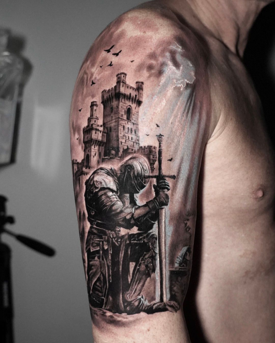 A black-and-grey tattoo on an upper arm featuring a knight kneeling before a sword in front of a stone castle.