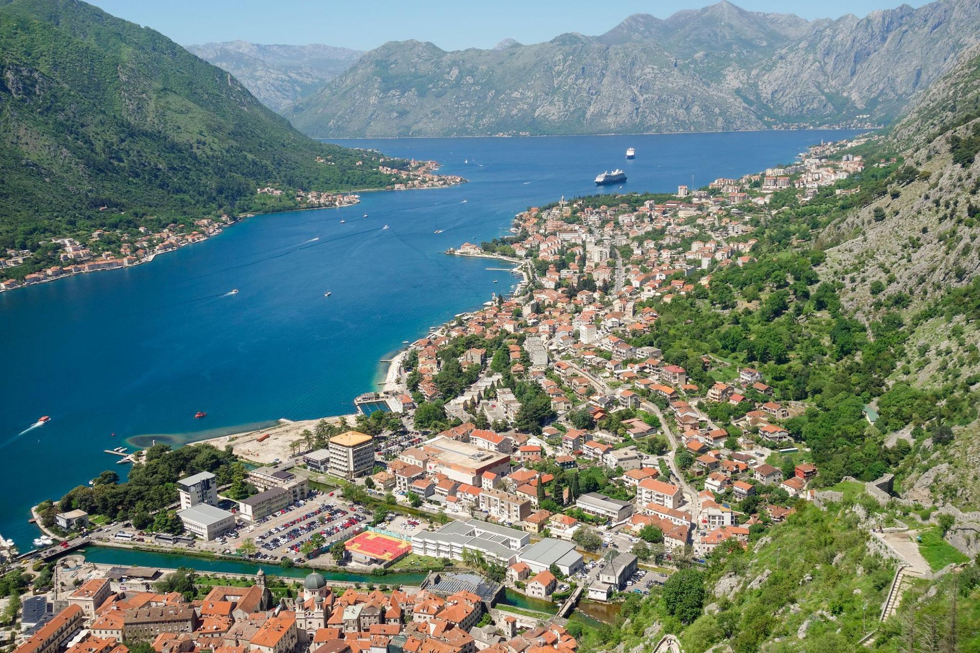 A coastal town beside a blue bay, with terracotta roofs and green mountains rising around it.