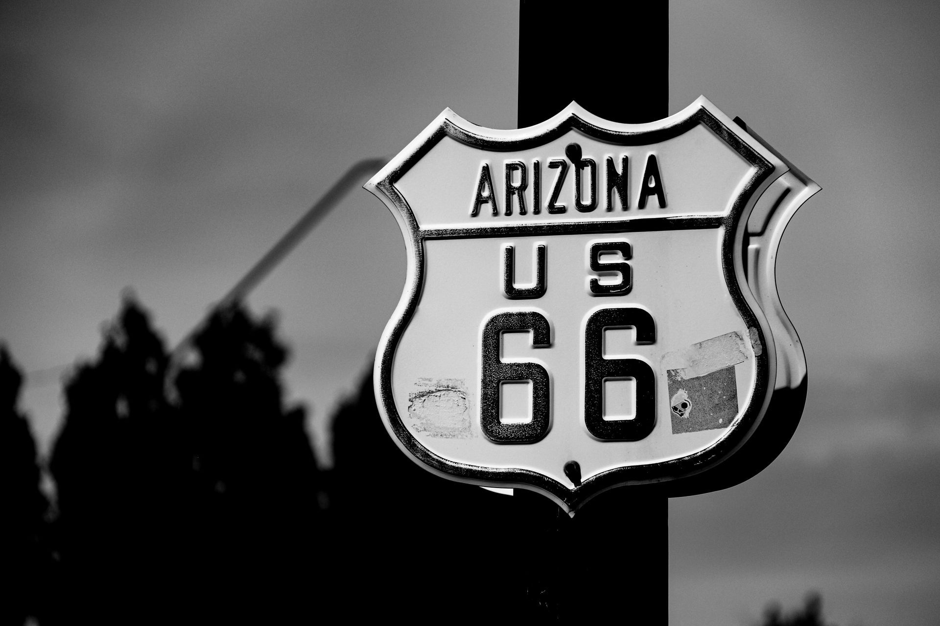 A black and white close-up of a shield-shaped Arizona US Route 66 road sign mounted on a post against a blurred background.