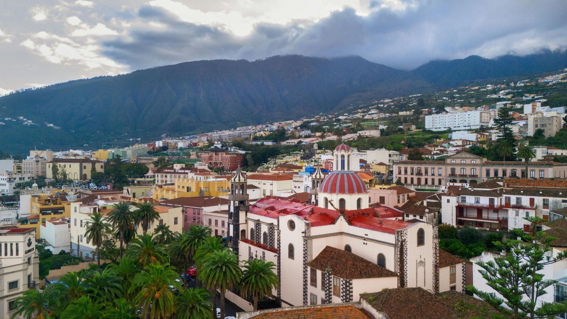Cityscape with colorful buildings, red-roofed church, and mountain backdrop under cloudy sky.