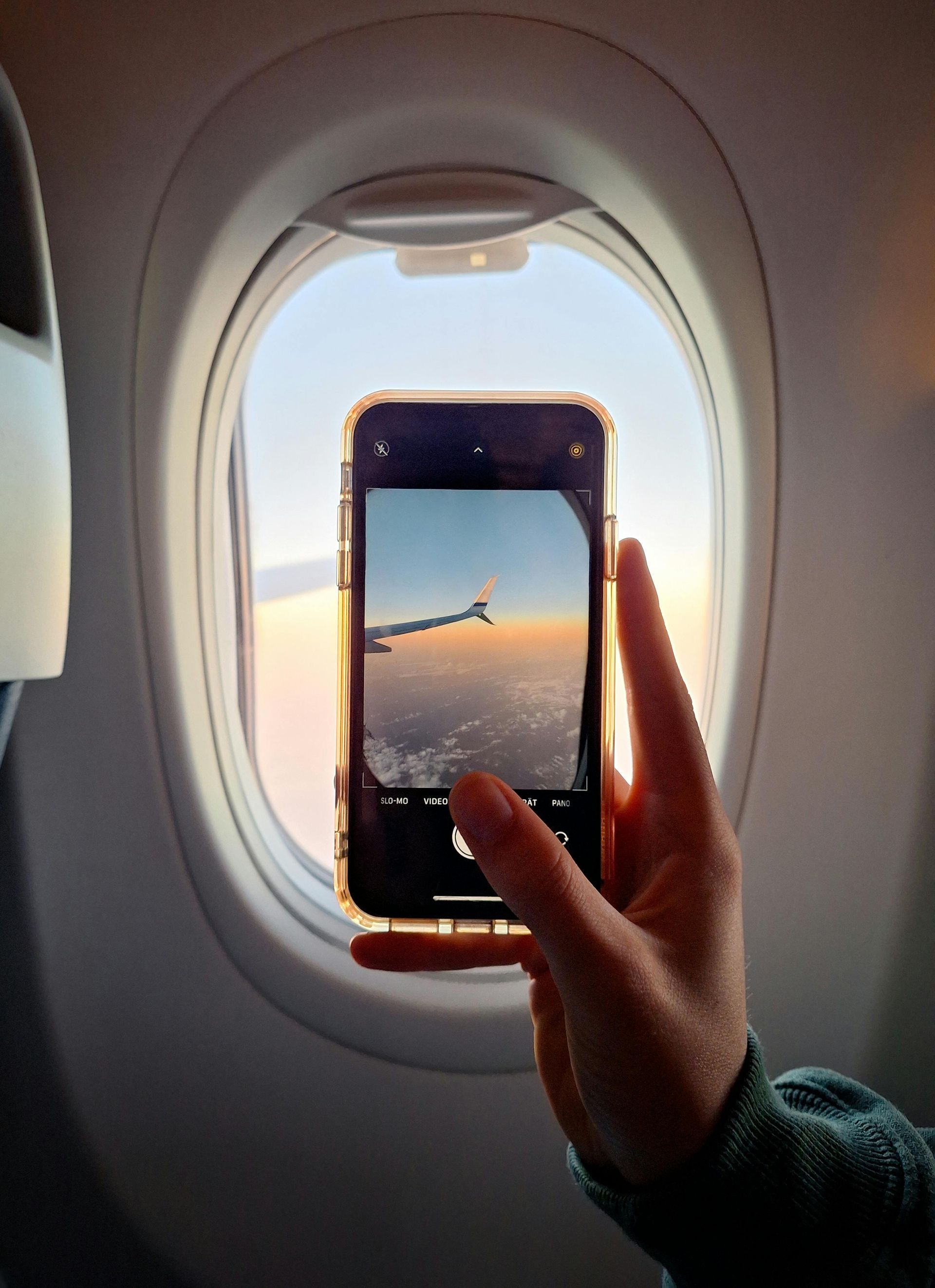 Hand holding a phone to an airplane window, showing a sunset sky and wing view.