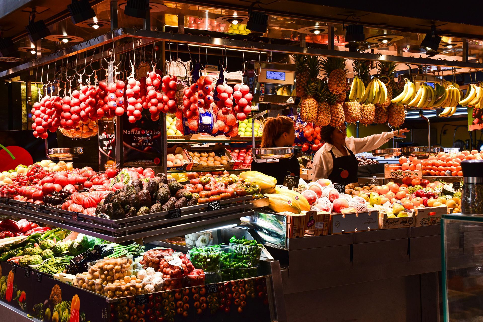 Fruit market stall with bunches of grapes, bananas, and other produce, vendor in background.