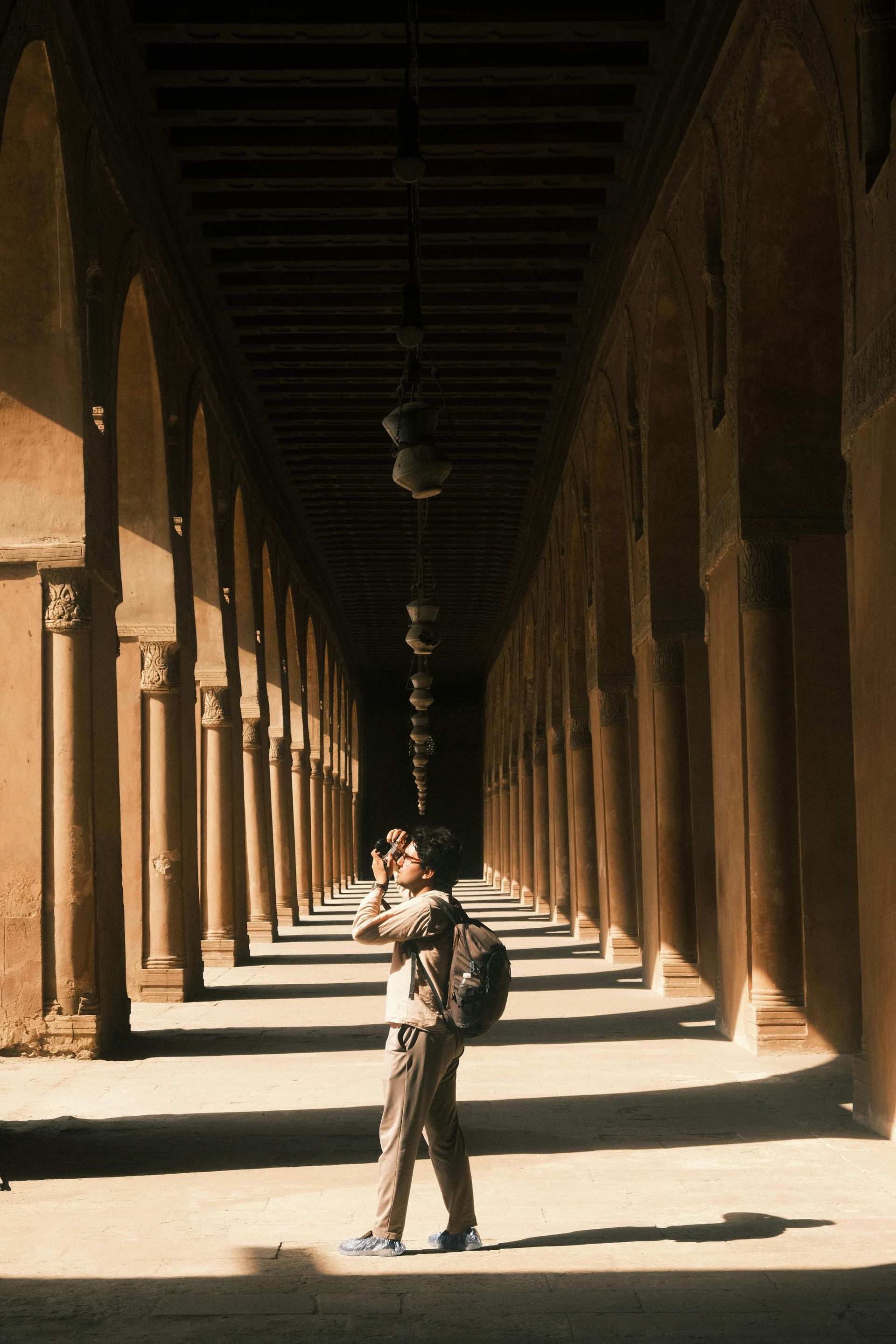 Person standing in a sunlit arched corridor with long repeating columns and strong shadows