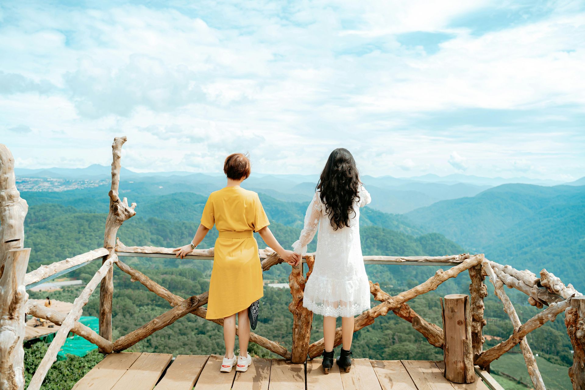 Two people stand on a wooden lookout, holding hands and facing a misty mountain valley.