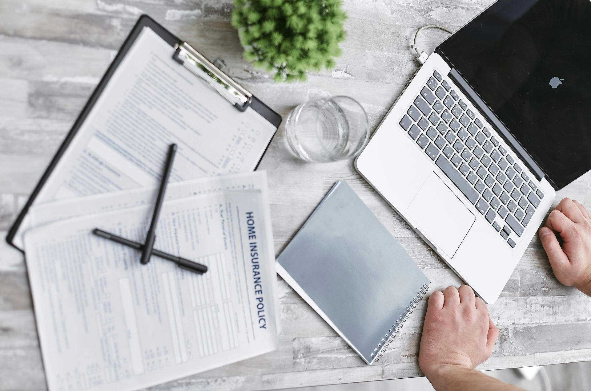 Workspace with laptop, clipboard, documents, pen, water glass, and hands.