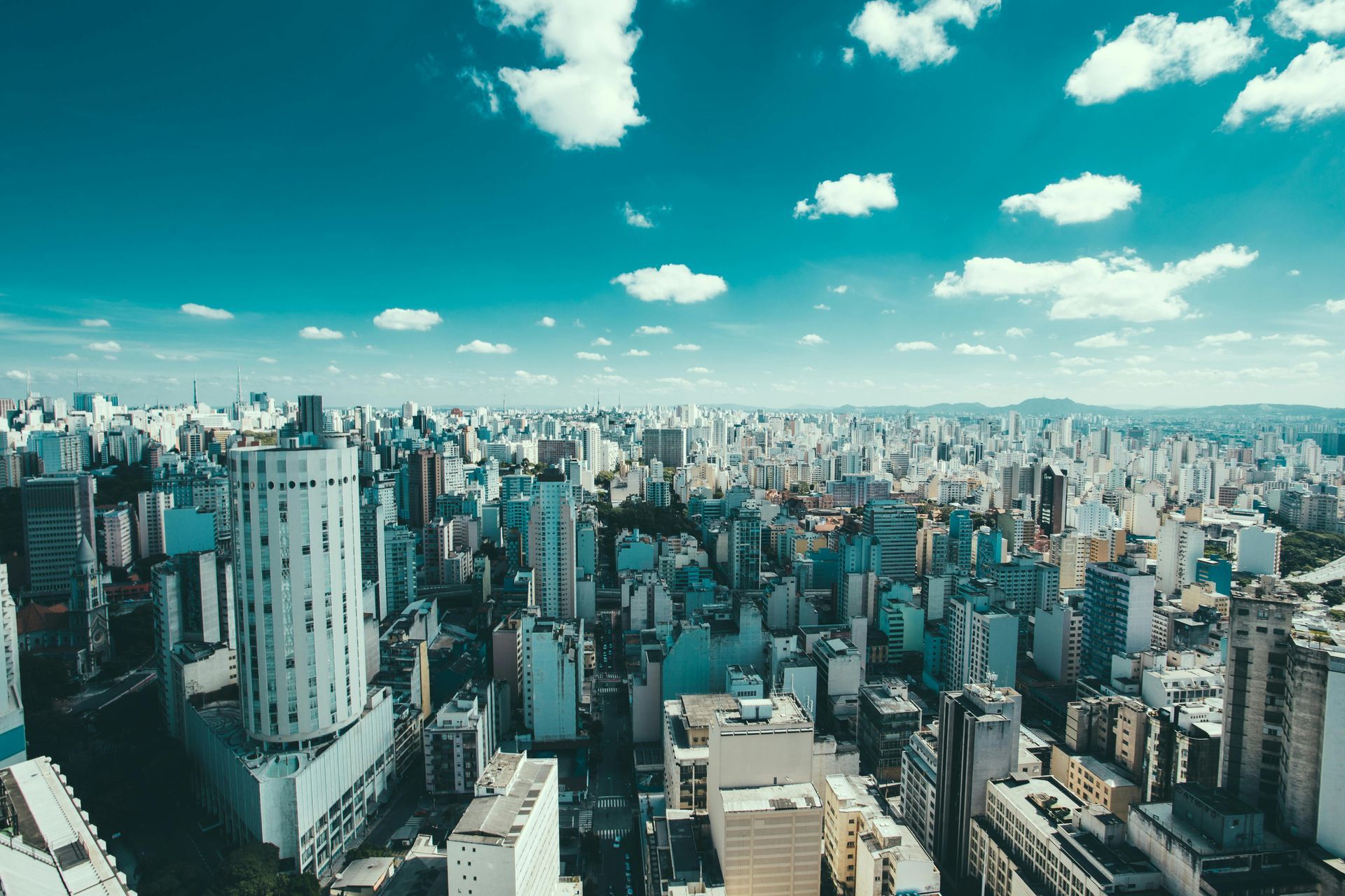 Aerial view of a densely populated city with numerous skyscrapers under a bright blue sky with fluffy clouds.
