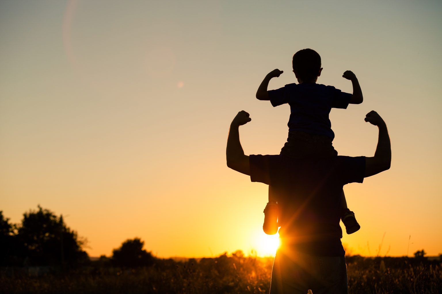 A silhouette of a child sitting on a parent's shoulders, both posing with arms flexed against a sunset background.