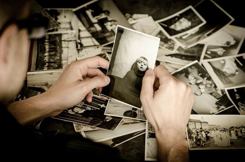 A person holds and looks at a small, black-and-white photograph, surrounded by a pile of various vintage photos.