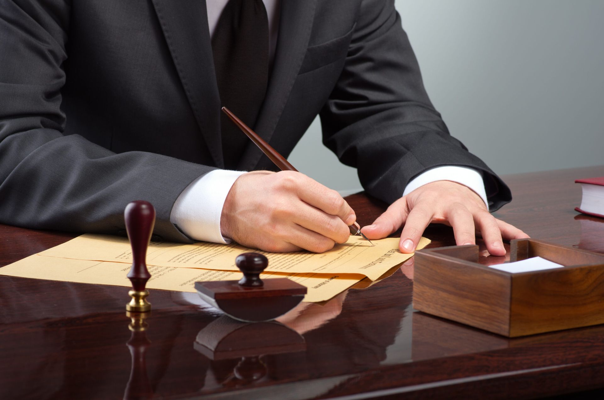 A person in a suit writes with a pen on paper at a desk, featuring a wax seal stamp, an ink pad, and a wooden box.