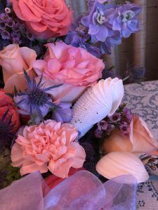 A floral arrangement with pink roses, purple thistles, blue delphiniums, and seashells accented with a purple ribbon.