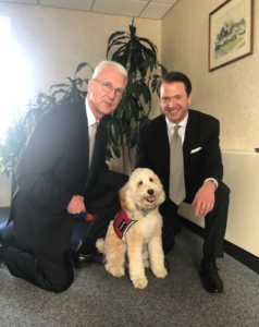 Two people in suits kneel on either side of a smiling, light-colored dog wearing a red harness in an office setting.