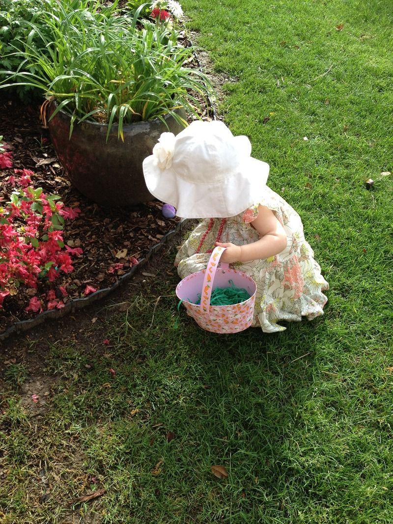 A child in a white hat and floral dress picks up items in a grass garden while holding a small patterned basket.