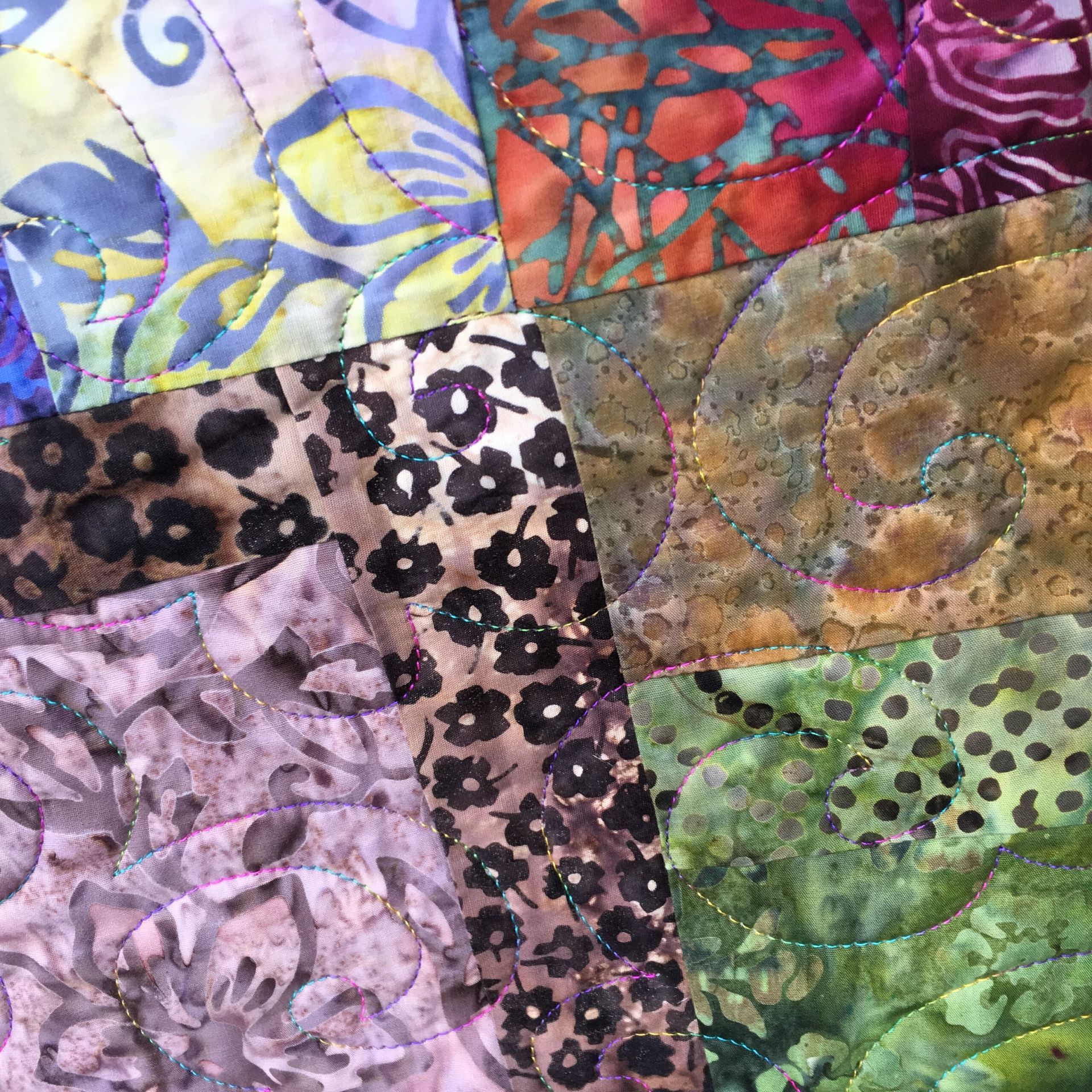 A close-up of a patchwork quilt featuring various batik fabric patterns in purple, green, orange, and floral designs.