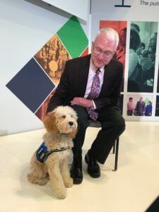 A man in a suit and patterned tie sits on a chair, smiling while resting his hand on a light-colored service dog.
