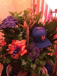 A floral basket featuring blue hydrangeas, orange ribbon, and a New York Mets baseball cap tucked among green foliage.