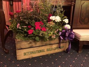 A wooden International Bulb Company crate overflowing with red, white, and purple flowers, adorned with a purple ribbon.