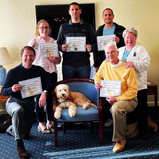 A group of people and a dog in a room holding certificates, with the dog seated in a blue chair.