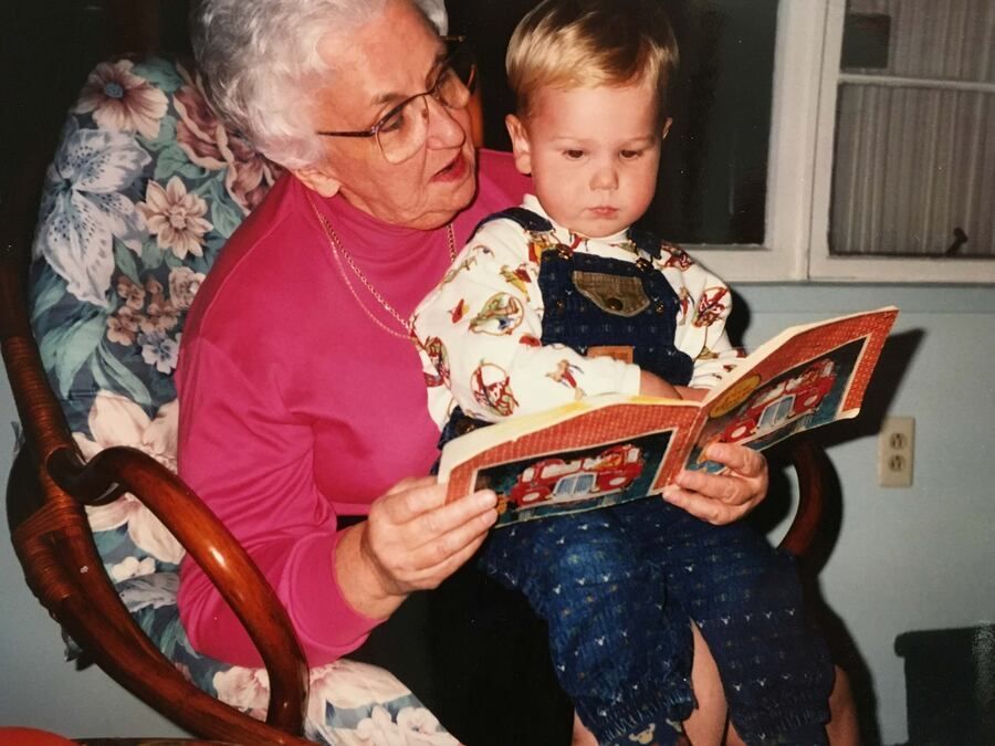 An older person in a pink shirt reads a book to a child in denim overalls while sitting in a patterned rocking chair.
