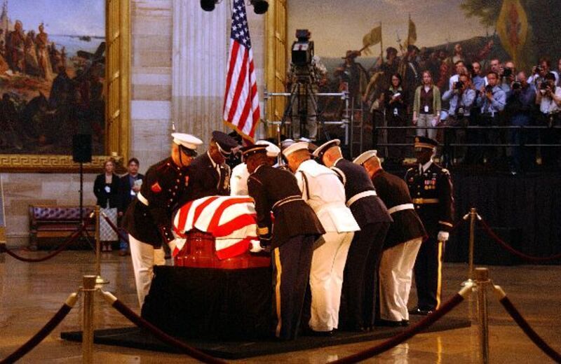 A casket draped in an American flag is being placed on a stand by uniformed military personnel inside a rotunda.