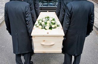 Four people in dark coats carry a light-colored wooden coffin topped with a floral arrangement toward a hearse.