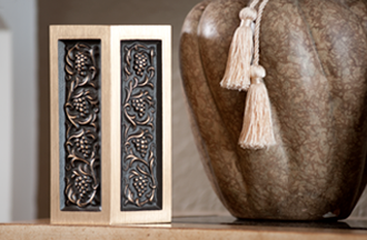 A rectangular urn with bronze-tone grape vine carvings sitting next to a brown, textured vase with hanging tassels.