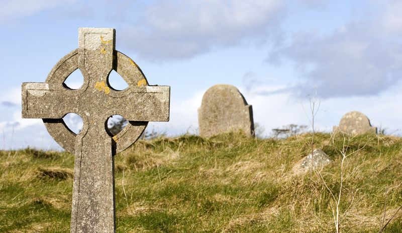 A weathered Celtic cross stands in a grassy cemetery against a cloudy blue sky, with other headstones visible nearby.